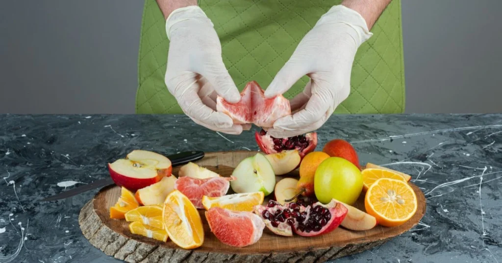 Hands wearing gloves preparing detox-friendly fruits like grapefruit, apples, pomegranates, and oranges on a wooden cutting board.