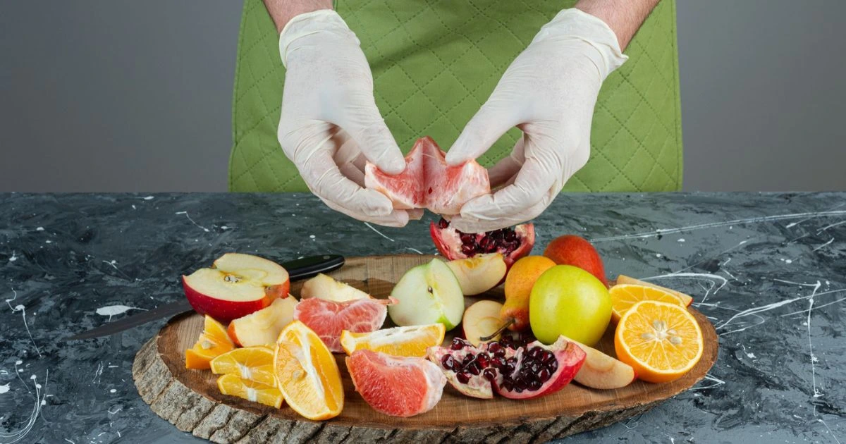 Hands wearing gloves preparing detox-friendly fruits like grapefruit, apples, pomegranates, and oranges on a wooden cutting board.