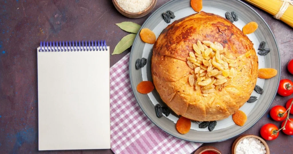 A round sweet potato bread topped with golden raisins, surrounded by dried apricots, raisins, and fresh ingredients on a rustic table.
